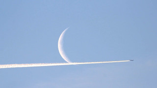 Widok błękitnego nieba z dużym, białym księżycem w tle, który przecina smuga lecącego samolotu. | A view of the blue sky with a large white moon in the background crossed by the plume of a flying plane.