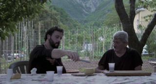 Dwóch mężczyzn siedzących przy stole, na świeżym powietrzu, jedzący ser i pijący wino. | Two men sitting at the table outdoors, eating cheese and drinking wine.