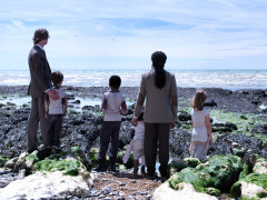 Zdjęcie przedstawiające bohaterów z dziećmi odwróconych plecami do widzów nad wodą. | A photo showing the characters with their children turned their backs to the viewers by the water.